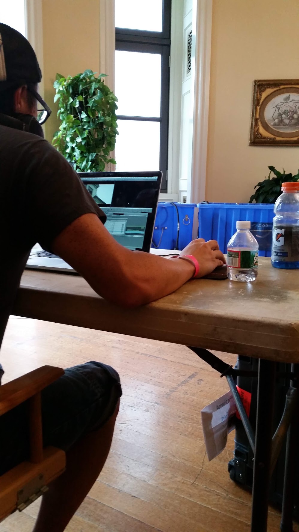 Justin editing on set at Oheka Castle — MacBook open on a wooden table, blue road cases, plants in the corner.